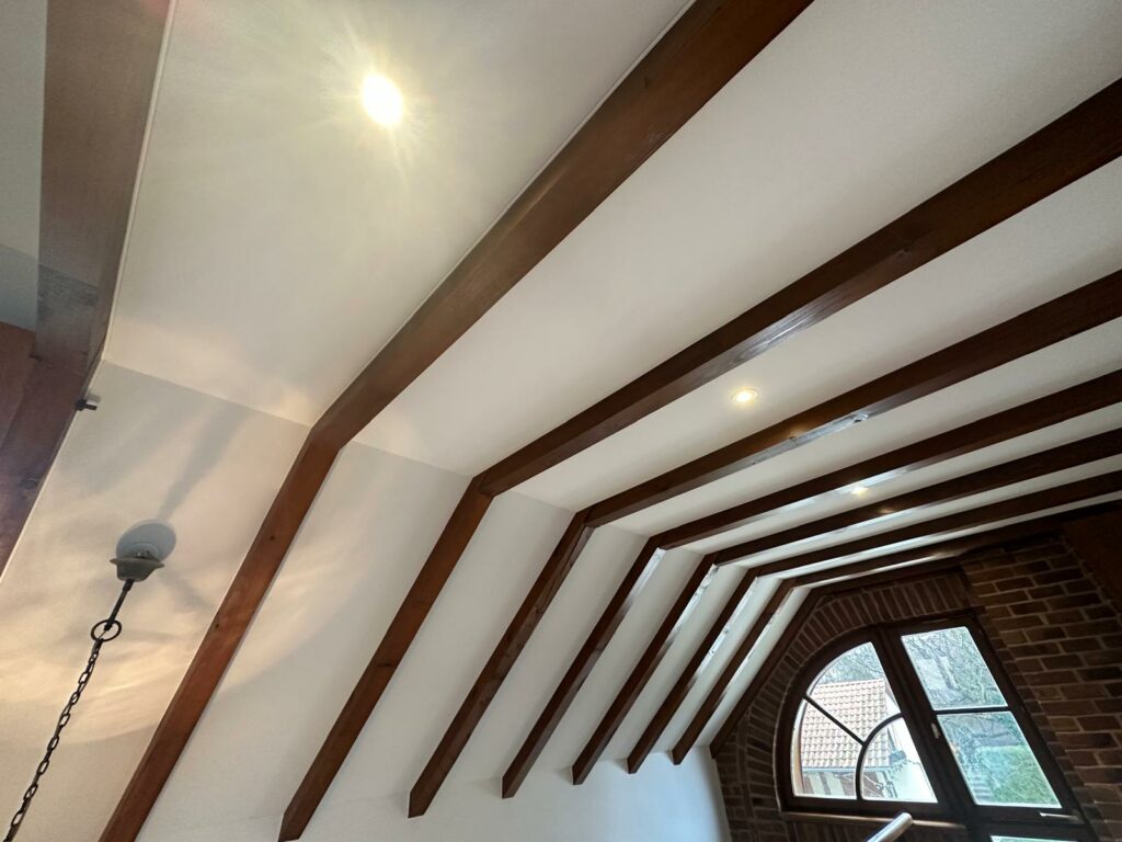 Plafond incliné avec poutres apparentes en bois et éclairage intégré, donnant sur une fenêtre arquée.
