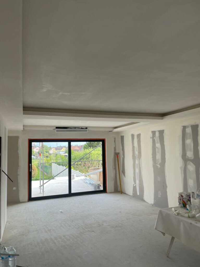 Salle en rénovation avec faux plafond et grandes fenêtres donnant sur un jardin.