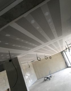 Plafond perforé en cours d'installation dans un espace ouvert en béton, avec des câbles suspendus.
