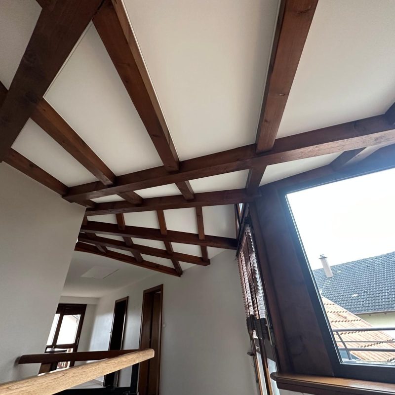 Plafond en bois avec poutres apparentes et panneaux intégrés, éclairant un couloir lumineux.