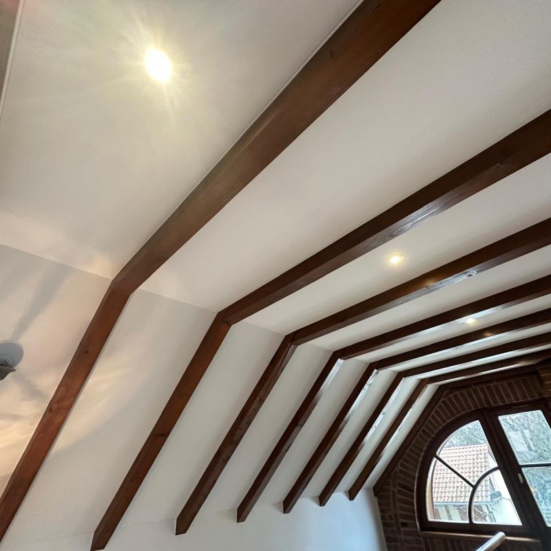 Plafond incliné avec poutres apparentes en bois et éclairage intégré, donnant sur une fenêtre arquée.