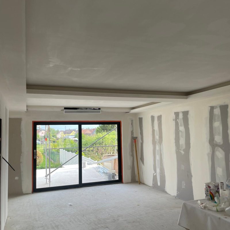 Salle en rénovation avec faux plafond et grandes fenêtres donnant sur un jardin.